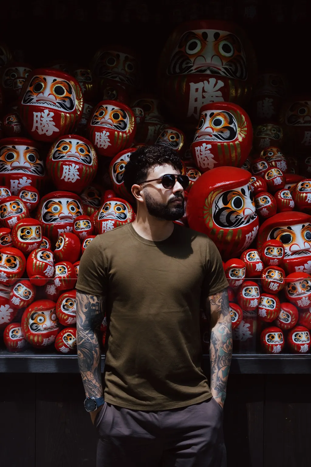 Retrato de Christofer Ramos diante de uma parede de daruma no Katsuo-ji, no Japão