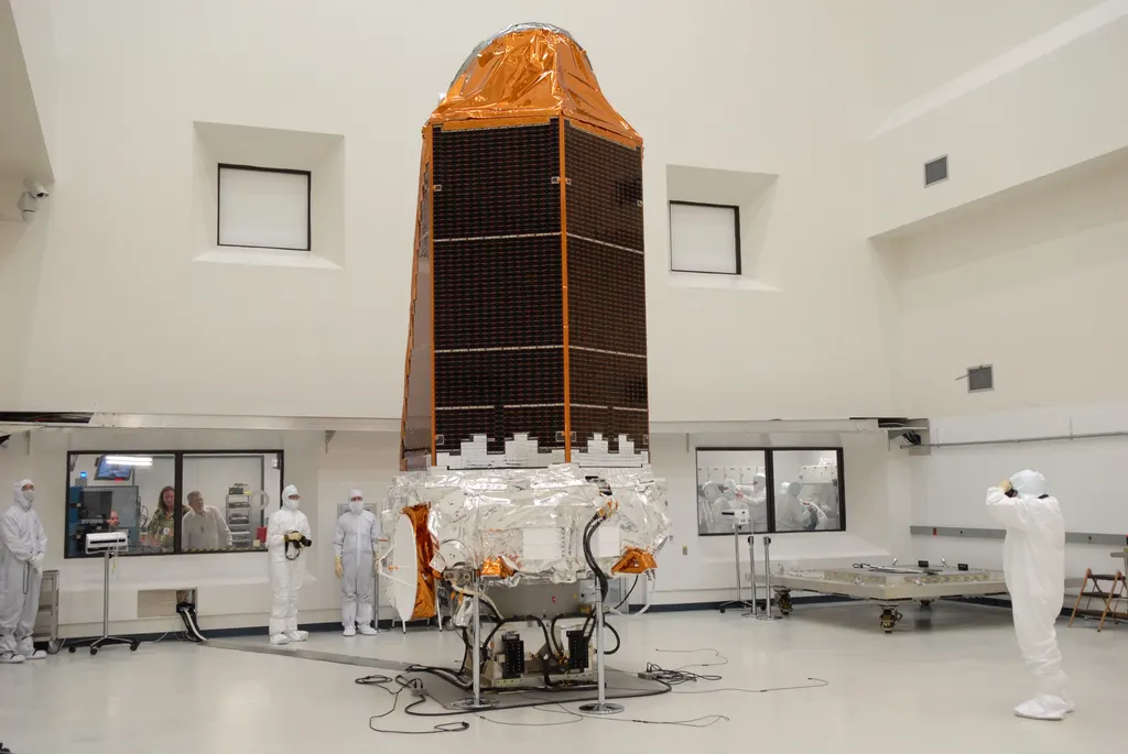 O telescópio espacial Kepler da NASA de pé numa sala limpa antes do lançamento: um corpo alto envolto em folha dourada e painéis solares escuros, com técnicos de macacão branco ao lado dando a escala.